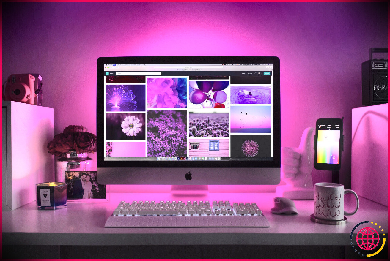 Un iMac est posé sur un bureau avec des lumières roses derrière lui et un iPhone à côté.  L'écran est allumé avec des images roses et violettes dessus
